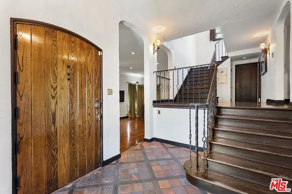 Foyer featuring a thick arched wooden front door, terracotta floor tiles, white plaster walls, and a dark wood staircase with wrought-iron railings.