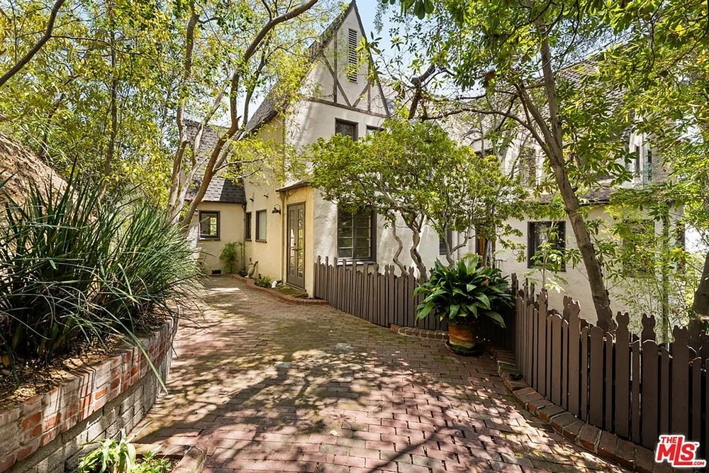 Brick-paved courtyard pathway leading along a classic Tudor-style home framed by mature trees and a wooden picket fence.