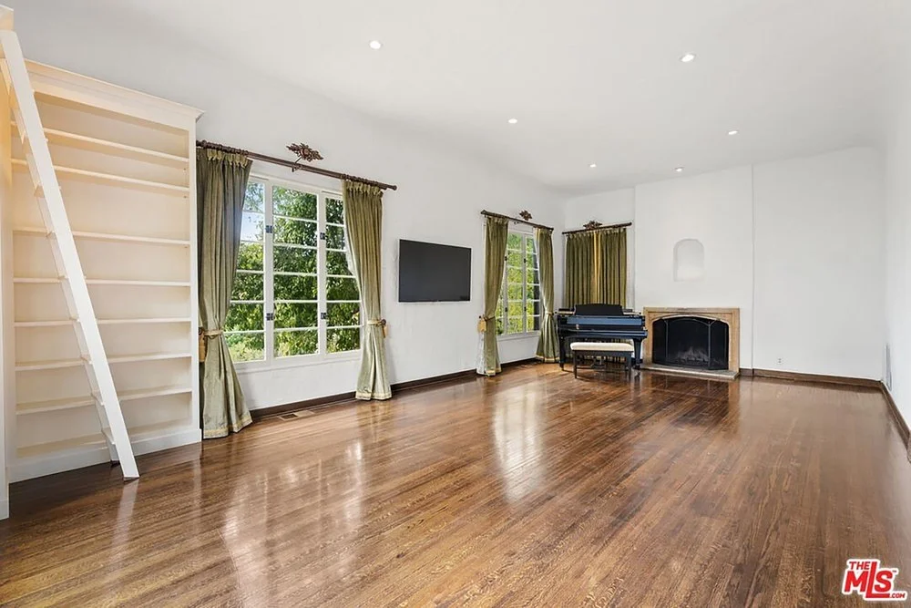 Spacious living area with dark hardwood floors, white library shelving, a black baby grand piano, olive green curtains, and a classic fireplace.